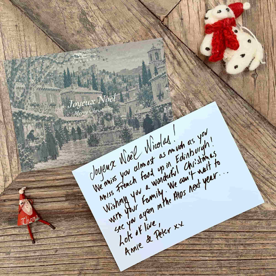 Tariette Joyeux Noël Christmas card with a handwritten festive message, photographed on rustic wood with Christmas decorations.