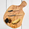 Wooden cutting board with cheese, fruit, and two knives on a white wooden surface