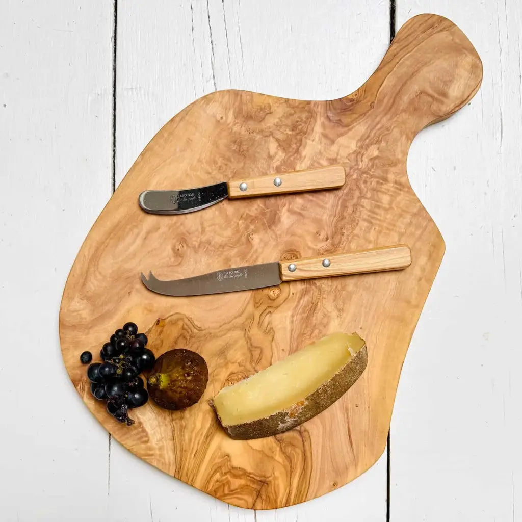Wooden cutting board with cheese, fruit, and two knives on a white wooden surface