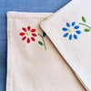Each napkin is hand-embroidered in one corner, four with a bright blue flower and four with a vivid red, both with fresh green leaves.